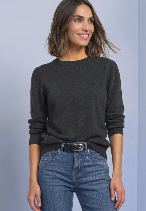 Une femme souriante en pull gris foncé et jean ceinturé, devant un fond clair.