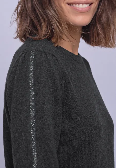 Une femme aux cheveux bruns mi-longs porte un pull gris foncé avec une bande argentée à l’épaule.