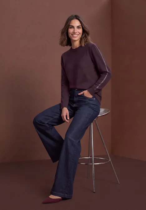 Femme souriante en pull violet et jean large, assise sur un tabouret devant un fond brun.