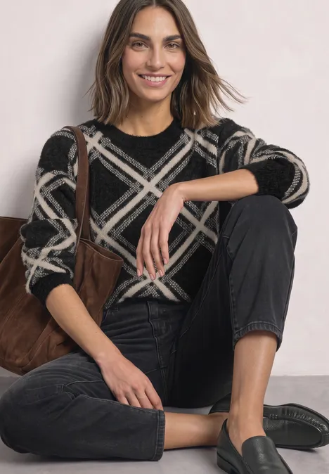 Femme assise au sol, souriante, porte un pull noir à carreaux beiges et un jean noir, chaussures style mocassins.