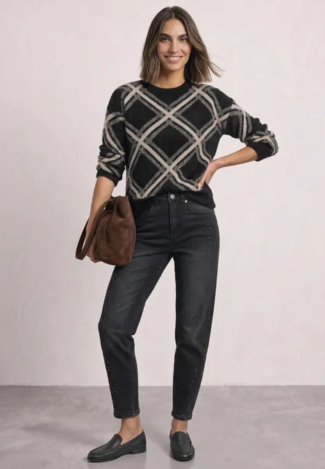 Femme en jean noir et pull à motifs, portant un sac marron et des mocassins devant un fond clair.