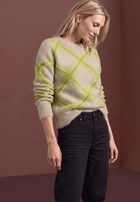 Pull à col rond et motif à carreaux clay sand Frau im hellbeigen Pullover mit grünen Rautenmustern und schwarzen Hosen vor braunem Hintergrund.