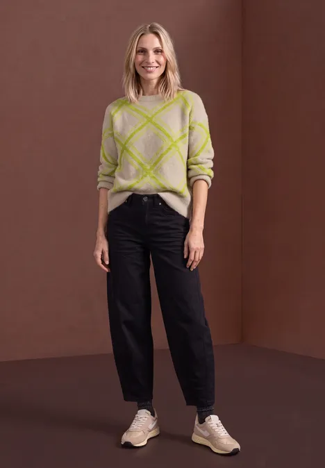 Pull à col rond et motif à carreaux clay sand Frau im beigen Karopullover, schwarzen Hose und hellen Sneakers vor braunem Hintergrund.