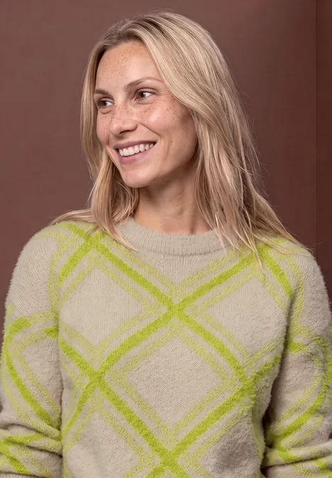 Pull à col rond et motif à carreaux clay sand Lächelnde blonde Frau in grobgestricktem Pullover mit grünen Rauten vor braunem Hintergrund.