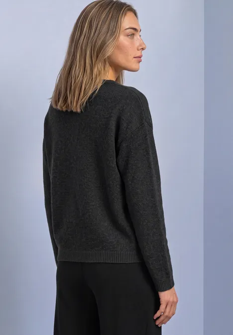 Femme de profil portant un pull et un pantalon noirs devant un mur clair.