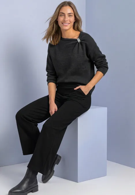 Une femme souriante en pull gris foncé, pantalon ample noir et bottines assise sur un cube clair.
