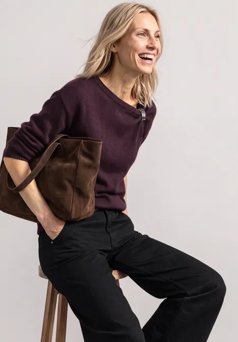 Pull à col bateau orné d'une broche berry plum mel. Eine lächelnde Frau in lila Pullover und schwarzer Hose hält eine braune Tasche und sitzt vor hellem Hintergrund.