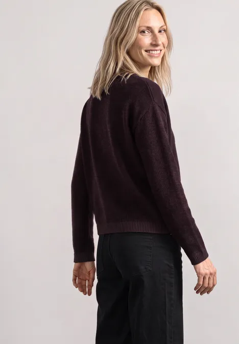 Pull à col bateau orné d'une broche berry plum mel. Frau mit blondem Haar trägt dunkelroten Pullover und schwarze Hose, schaut über die Schulter und lächelt.