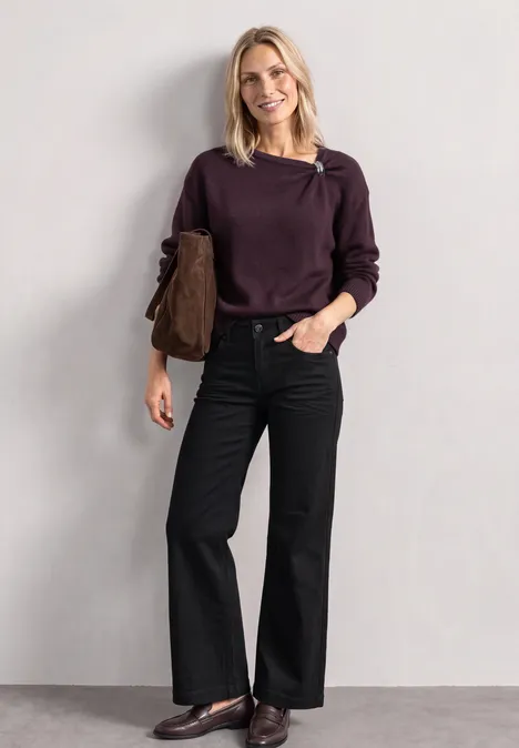 Pull à col bateau orné d'une broche berry plum mel. Eine lächelnde Frau mit blondem Haar trägt einen lila Pullover, schwarze weite Hosen und eine braune Tasche.