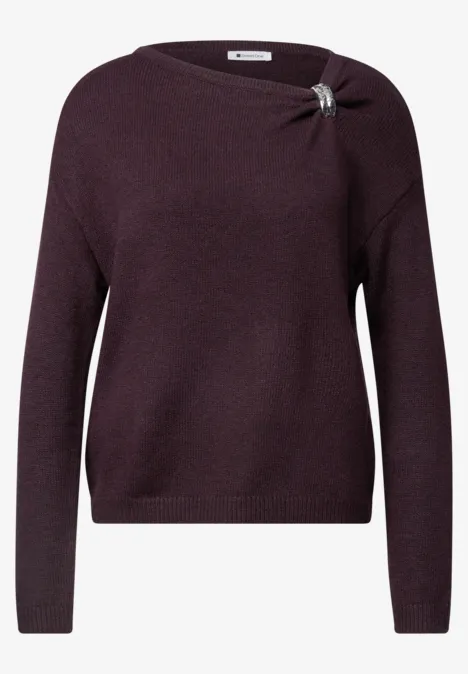 Pull à col bateau orné d'une broche berry plum mel. Violetter Pullover mit langen Ärmeln, Knotendetail und silberner Brosche an der Schulter.