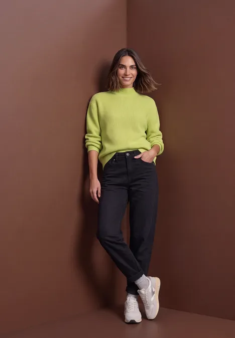 Pullover im Melange-Look mit Turtle Neck apple lime mel. Frau in hellgrünem Pullover, schwarzen Jeans und weißen Turnschuhen vor braunem Hintergrund, lächelnd.