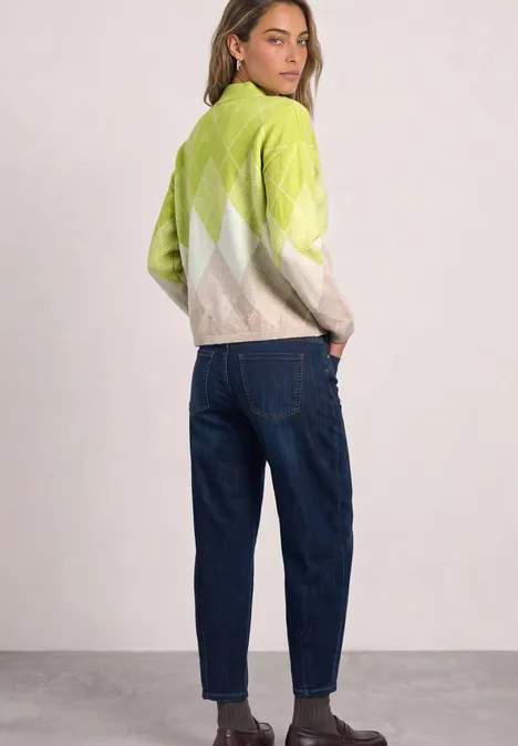 Frau in grün-weißem Argyle-Pullover, blauen Jeans und braunen Stiefeletten, schaut über die Schulter.