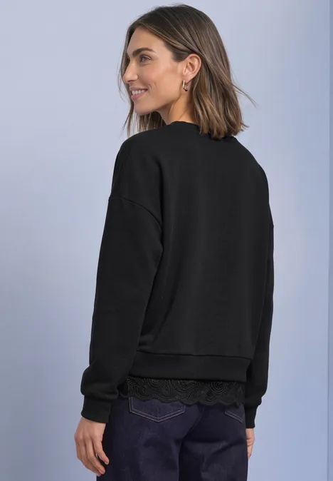 Loose Fit Sweatshirt mit Spitzenkante Black Frau in lockerem schwarzen Sweatshirt vor hellem Hintergrund, lächelnd zur Seite blickend.