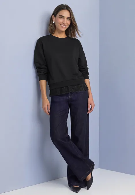 Loose Fit Sweatshirt mit Spitzenkante Black Frau in schwarzem Pullover und weitem Jeans, lächelt vor hellblauem Hintergrund.