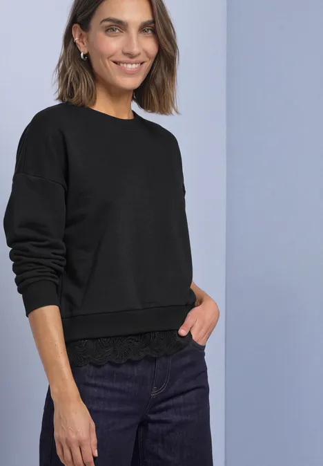 Loose Fit Sweatshirt mit Spitzenkante Black Frau mit kurzem braunem Haar trägt schwarzen Pullover mit Spitzenbund und dunklen Jeans, sie lächelt.