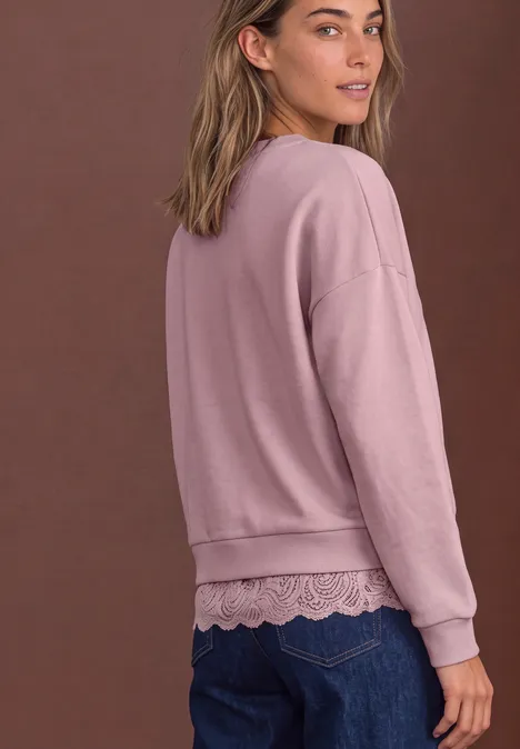 Loose Fit Sweatshirt mit Spitzenkante quiet rose Frau von hinten in rosa Sweatshirt mit Spitzenabschluss über blauen Jeans vor braunem Hintergrund.