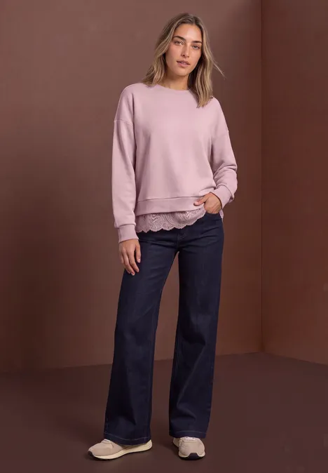 Loose Fit Sweatshirt mit Spitzenkante quiet rose Frau in rosa Sweatshirt und weiten Jeans steht vor braunem Hintergrund, lässige Pose mit Händen in Taschen.