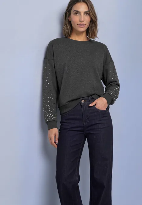Loose Fit Sweatshirt mit Nieten armor grey mel. Frau trägt dunkles Sweatshirt mit Nieten an den Ärmeln und dunkle Jeans, steht vor hellem Hintergrund.