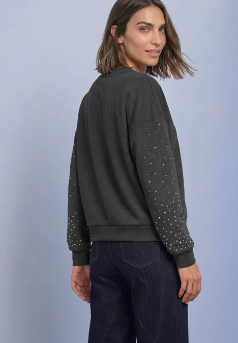 Loose Fit Sweatshirt mit Nieten armor grey mel. Frau schaut über die Schulter, trägt dunkelgrauen Pullover mit Nietenärmeln und Jeans vor hellem Hintergrund.