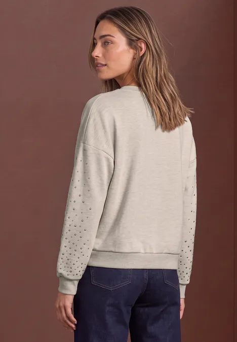 Sweatshirt met studs – Loose Fit clay sand mel. Rückansicht einer Frau in grauem Sweatshirt mit Lochmuster an den Ärmeln und blauen Jeans vor braunem Hintergrund.