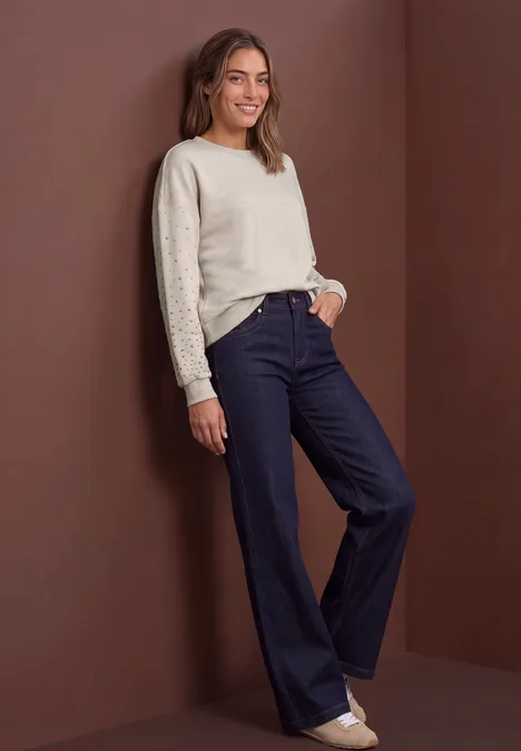 Sweatshirt met studs – Loose Fit clay sand mel. Frau in cremefarbenem Pullover und dunklen, weiten Jeans lehnt lässig an einer Wand.