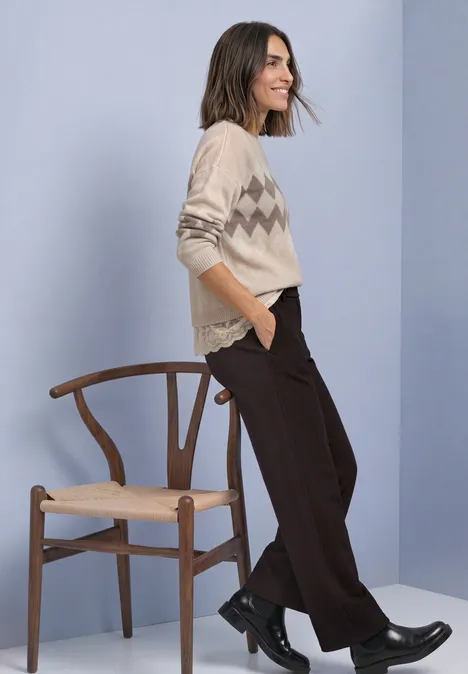 Pullover mit Jacquardmuster und Spitze clay sand mel. Frau in hellbeigem Zickzack-Pullover lehnt lässig am Holzstuhl, trägt dunkle Hose und schwarze Stiefel.
