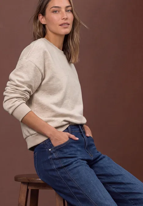 Eine Frau in beigem Pullover und blauen Jeans lehnt lässig auf einem Hocker vor braunem Hintergrund.