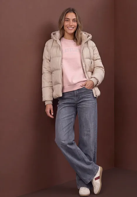 Frau in heller Steppjacke, rosa Pullover und blauen Jeans steht entspannt und lächelt.