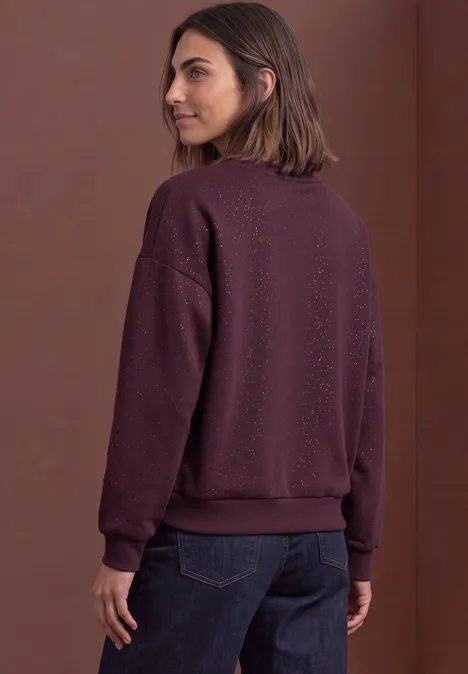 Frau mit mittellangen braunen Haaren in lila Sweatshirt und dunklen Jeans, seitlich von hinten aufgenommen.