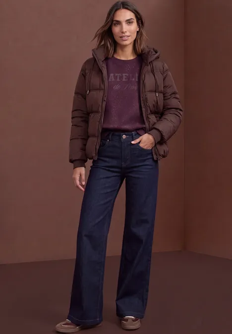 Eine Frau steht vor braunem Hintergrund und trägt eine dunkelbraune Daunenjacke, weite Jeans und Pullover.