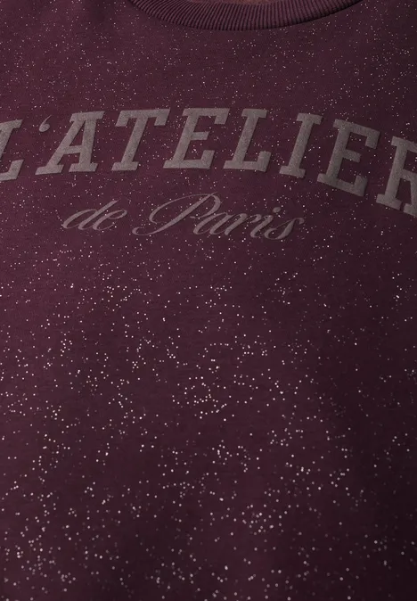 Nahaufnahme eines bordeauxroten Pullovers mit Schriftzug 'L'ATELIER de Paris' und weißen Sprenkeln.