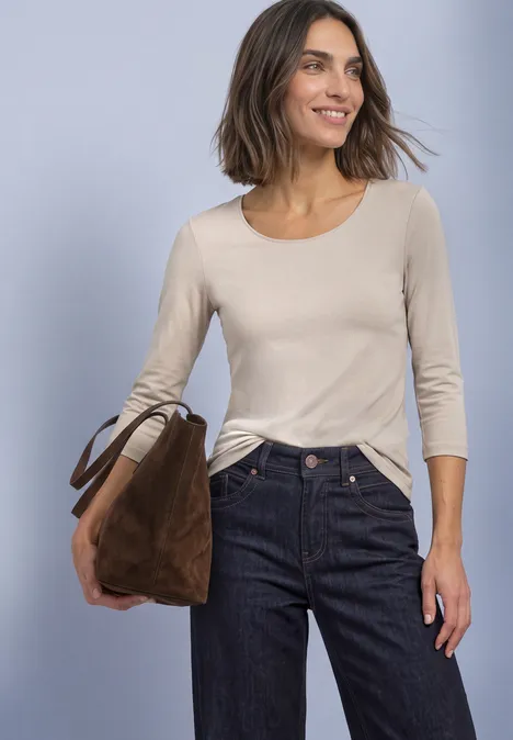 Eine Frau trägt ein beiges Langarmshirt, dunkle Jeans und hält eine braune Tasche.