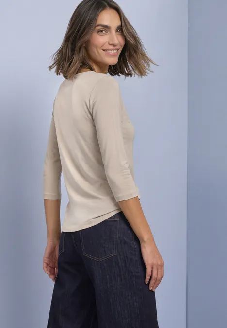 Frau mit beigefarbenem 3/4-Ärmel-Shirt und dunklen Jeans blickt über die Schulter und lächelt.