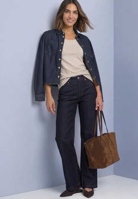 Frau trägt Jeansjacke, beiges Top, weite dunkle Jeans, schwarze Schuhe und braune Tasche vor hellem Hintergrund.