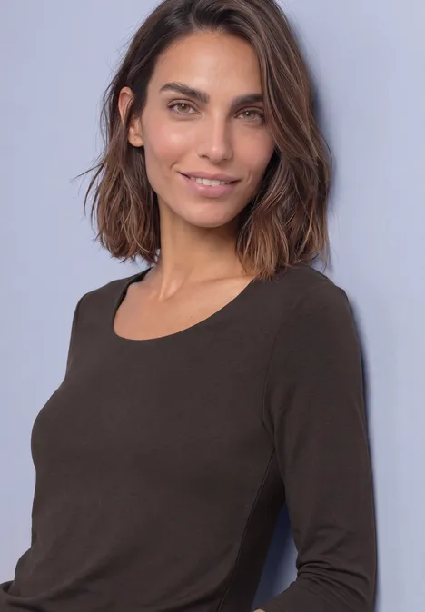 Vrouw met halflang bruin haar draagt een effen donkerbruin shirt met lange mouwen en glimlacht zacht.