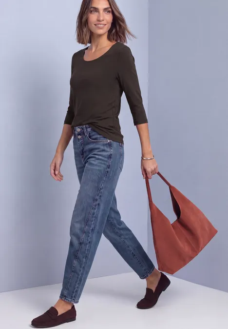 Een vrouw met een zwart 3/4-top, blauwe jeans en een bruine schoudertas voor een neutrale achtergrond.