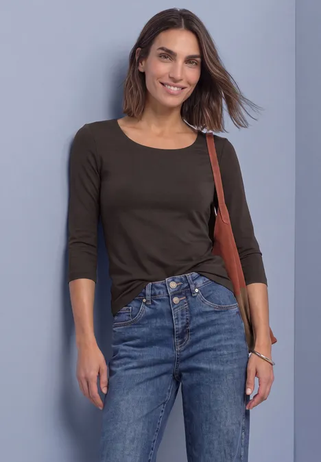 Bruine vrouw in zwarte top met 3/4-mouw en blauwe jeans glimlachend tegen een muur.