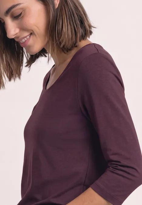 Une femme de profil souriante portant un t-shirt bordeaux à manches trois-quarts.