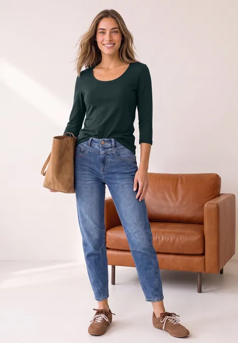 Junge Frau steht in einem hellen Raum, trägt grüne Bluse, blaue Jeans, braune Schuhe und hält eine Tasche.