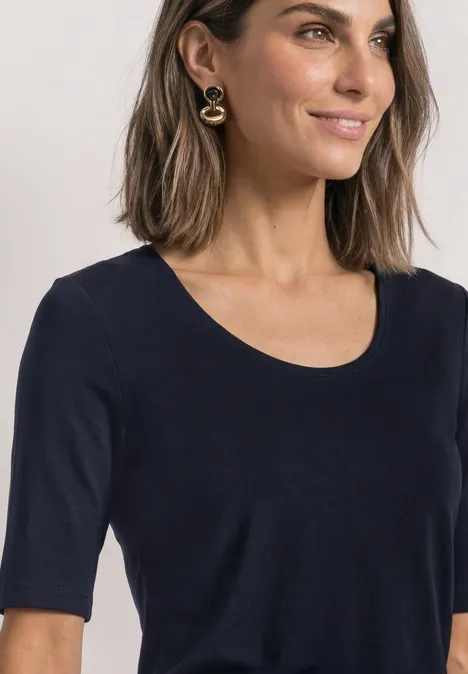 Frau mit schulterlangen Haaren, trägt schlichtes schwarzes Shirt mit Rundhals, halblangen Ärmeln und goldenen Ohrringen.