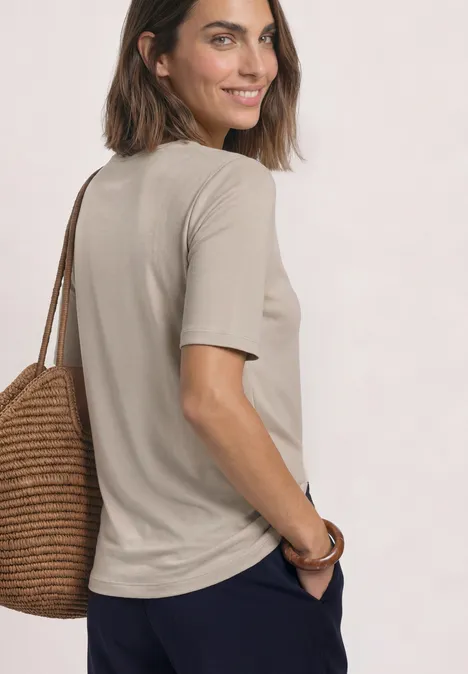 Frau im beigen T-Shirt mit Strohtasche, schaut lächelnd über die Schulter vor hellem Hintergrund.