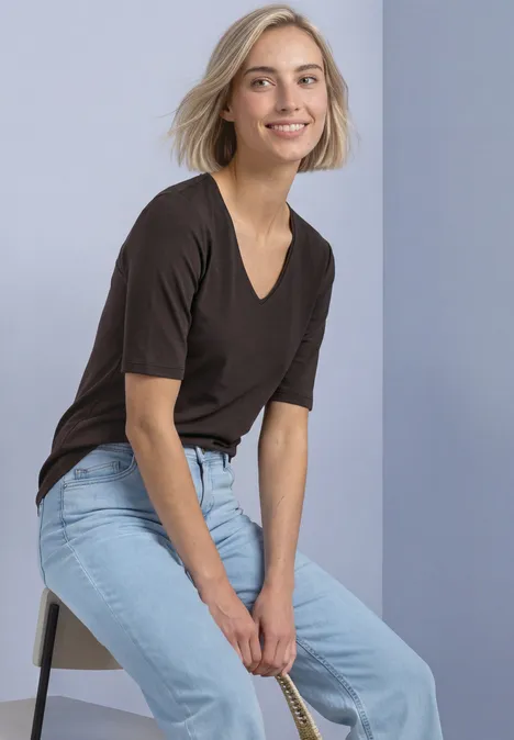 Een blondine zit op een krukje, glimlacht en draagt een zwart T-shirt en lichtblauwe jeans voor neutrale achtergrond.