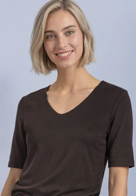 Een blonde vrouw met kort haar draagt een bruin shirt en lacht voor een effen achtergrond.