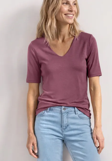 Frau im kurzärmligen V-Ausschnittshirt in Rosé und hellblauer Jeans vor weißer Wand.