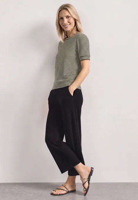 Frau in grauem Kurzarmshirt, schwarzen Hosen und Sandalen vor heller Wand.