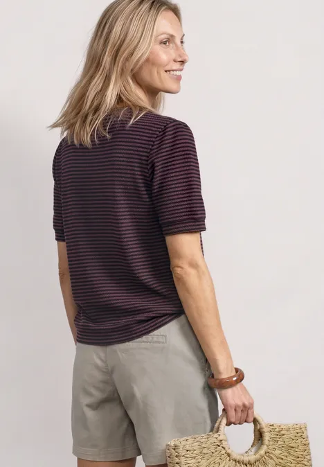Frau von hinten in gestreiftem T-Shirt und beigen Shorts, hält runde Strohtasche.