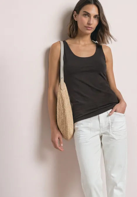 Basic top met ronde hals in effen kleur black coffee Jonge vrouw voor neutrale achtergrond, draagt een zwart topje, witte broek en een strotas.