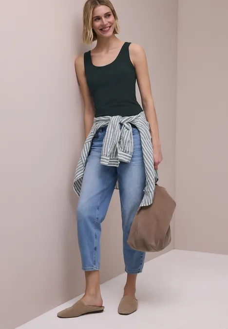 Frau in schwarzem Top, Jeans, Hemd um die Hüfte gebunden, trägt Tasche und Slipper.