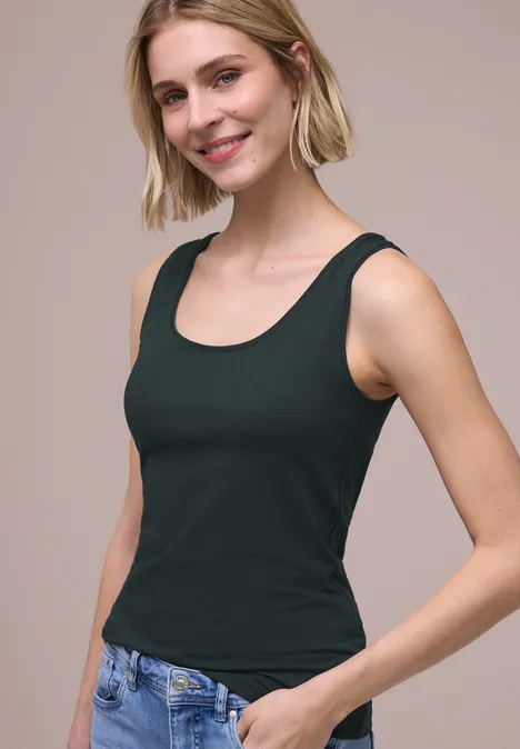 Eine Person mit kurzem blondem Haar trägt ein schwarzes ärmelloses Top und blaue Jeans.