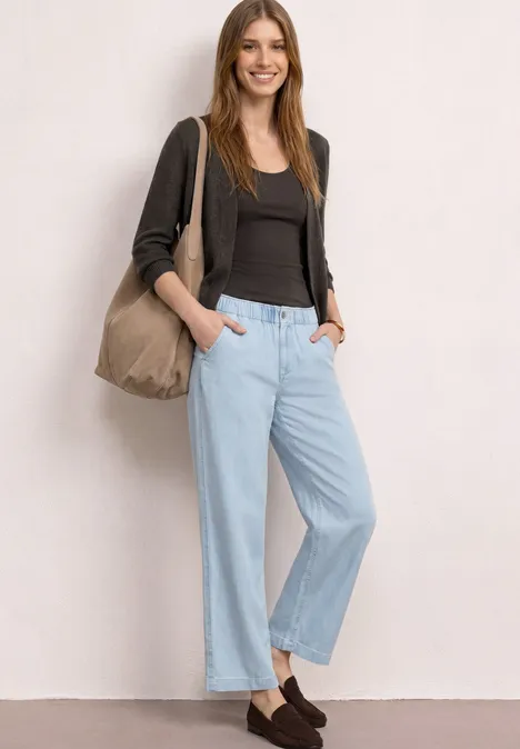 Frau in lässigem Outfit: schwarze Bluse, graue Strickjacke, helle, weite Jeans und braune Loafers mit Beuteltasche.